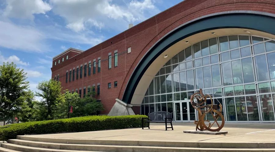 A long rectangular building with an arch of glass windows in the center on UofL's campus. Out front is a metal sculpture and the day is sunny with clouds in the sky.
